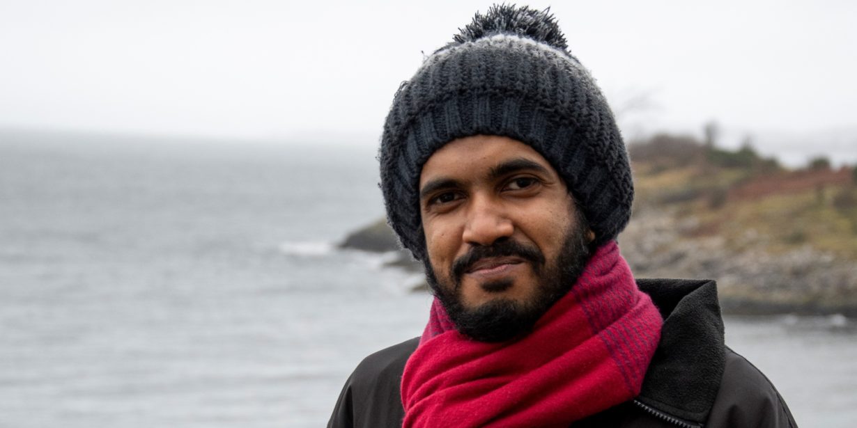 Sri Lankan student Faazil who is studying on the ACES+ masters programme poses for the camera in hat and scarf on the SAMS campus balcony on a cold winter afternoon.