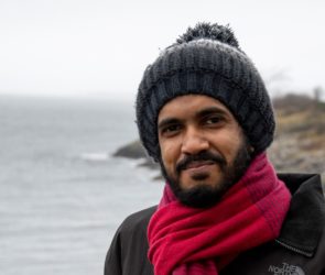 Sri Lankan student Faazil who is studying on the ACES+ masters programme poses for the camera in hat and scarf on the SAMS campus balcony on a cold winter afternoon.