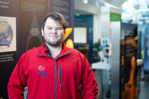 2017 SAMS UHI alumni Blair Watson poses for the camera in his red Dynamic Earth uniform jacket whilst on a recent work trip back to his old uni campus at SAMS UHI in Oban.