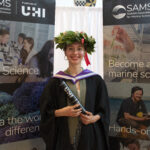 Italian student Ilaria Stolberg stands by two UHI banners holding her graduation scroll in graduation robes after she graduates from SAMS' BSc (Hons) Marine Science course wearing a wreath on her head with green foliage and red ribbon, traditionally worn in Italy to celebrate success and victory.