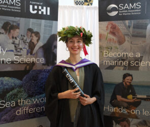 Italian student Ilaria Stolberg stands by two UHI banners holding her graduation scroll in graduation robes after she graduates from SAMS' BSc (Hons) Marine Science course wearing a wreath on her head with green foliage and red ribbon, traditionally worn in Italy to celebrate success and victory.