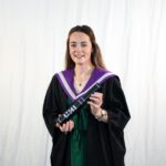 Kayleigh Roche poses for camera in studio at Graduation holding her scroll and wearing traditional graduation gown featuring purple colours.