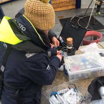 Kayleigh dressed in weatherproof sailing jacket and wooly hat works with samples on board the SAMS research vessel Seol Mara.