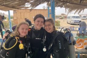 Isadora, far right, poses with two other female students in their wetsuits and scuba diving gear in the outdoor prep area, smiling and happy.