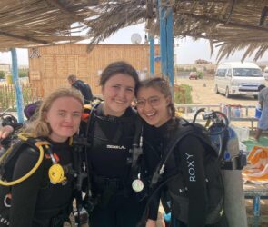 Isadora, far right, poses with two other female students in their wetsuits and scuba diving gear in the outdoor prep area, smiling and happy.