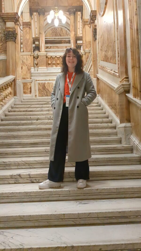 SAMS Algal Biotech student Evie Whyte stands smiling on a grand marble staircase wearing her event landyard at the IBioIC annual conference at Glasgow's City Chambers in March 2024.