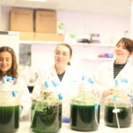 Image credit: Phyco Studio. Perle (left), Kristin (centre) and Evie (right) smile in white lab coats next to four large carboys of green algae during their lab time at Cambridge Algal Innovation Centre. In this picture they are learning different ways of growing and harvesting algae in larger settings.