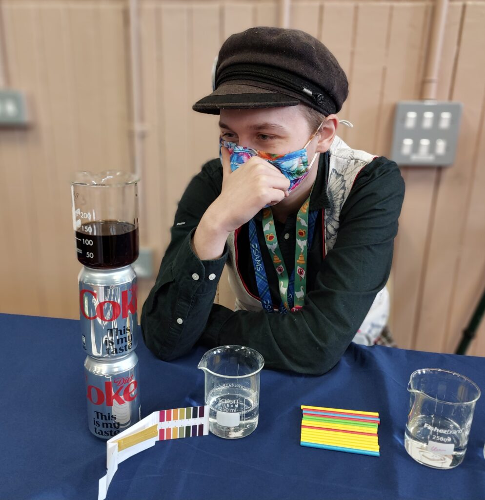 Artemis sits at a display table with cans of coke and ph strips ready to speak to children about ocean acidification.